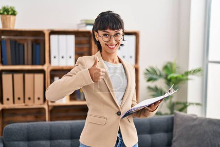 Photo for Young hispanic woman working at consultation office smiling happy and positive, thumb up doing excellent and approval sign - Royalty Free Image