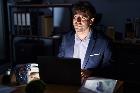 Photo for Hispanic young man working at the office at night smiling looking to the side and staring away thinking. - Royalty Free Image
