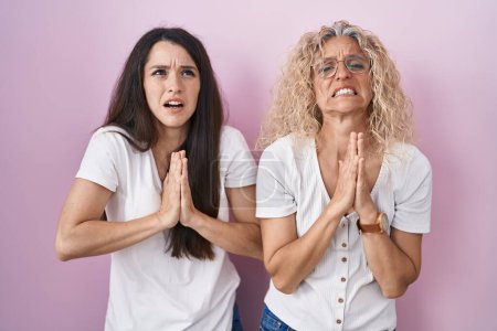Photo for Mother and daughter standing together over pink background begging and praying with hands together with hope expression on face very emotional and worried. begging. - Royalty Free Image