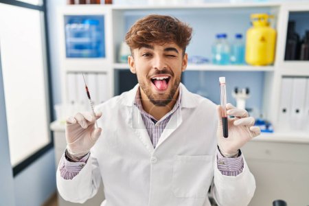 Foto de Hombre árabe con barba trabajando en laboratorio científico sosteniendo muestra de sangre sacando la lengua feliz con expresión divertida. - Imagen libre de derechos
