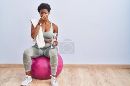 Téléchargez les photos : Femme afro-américaine portant des vêtements de sport assis sur une balle de pilates sentant quelque chose de puant et dégoûtant, odeur intolérable, retenant son souffle avec les doigts sur le nez. mauvaise odeur - en image libre de droit