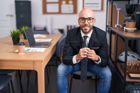 Téléchargez les photos : Jeune homme chauve travailleur d'affaires souriant confiant assis sur la table au bureau - en image libre de droit