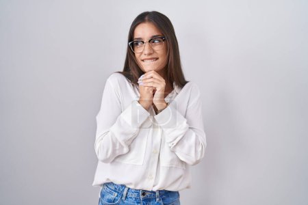Téléchargez les photos : Jeune femme brune portant des lunettes riant nerveux et excité avec les mains sur le menton regardant vers le côté - en image libre de droit