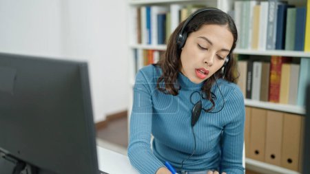 Téléchargez les photos : Jeune belle étudiante hispanique utilisant l'ordinateur prenant des notes portant des écouteurs à l'université salle de classe - en image libre de droit