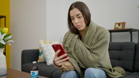 Photo for Young beautiful hispanic woman covering with blanket for cold using smartphone at home - Royalty Free Image