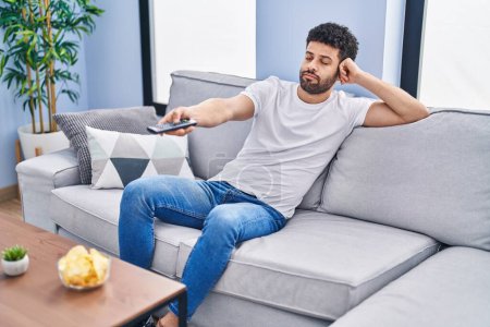 Téléchargez les photos : Jeune homme arabe regarder un film assis sur le canapé à la maison - en image libre de droit