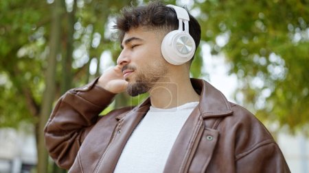 Téléchargez les photos : Jeune homme arabe écoutant de la musique avec une expression détendue au parc - en image libre de droit