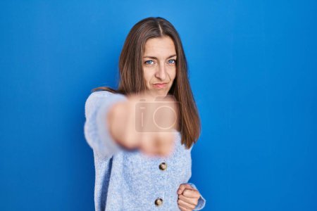 Photo for Young woman standing over blue background punching fist to fight, aggressive and angry attack, threat and violence - Royalty Free Image