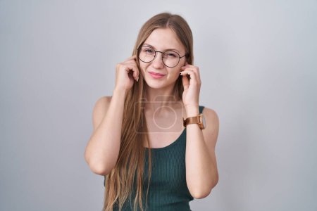 Téléchargez les photos : Jeune femme caucasienne debout sur fond blanc couvrant les oreilles avec des doigts avec une expression agacée pour le bruit de la musique forte. concept sourd. - en image libre de droit