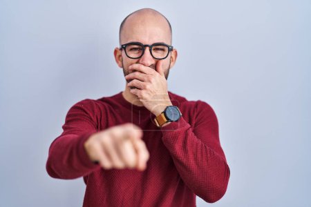 Photo for Young bald man with beard standing over white background wearing glasses laughing at you, pointing finger to the camera with hand over mouth, shame expression - Royalty Free Image
