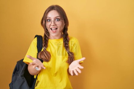 Téléchargez les photos : Jeune femme caucasienne portant un sac à dos étudiant sur fond jaune souriant gai à bras ouverts comme accueil amical, salutations positives et confiantes - en image libre de droit