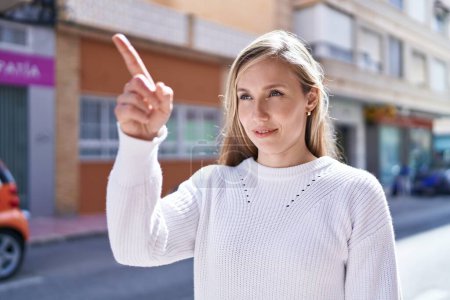 Téléchargez les photos : Jeune femme blonde souriante confiante pointant du doigt la rue - en image libre de droit