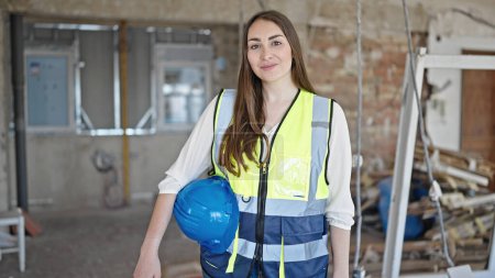 Téléchargez les photos : Jeune belle hispanique femme constructeur souriant confiant tenant hardhat sur le chantier de construction - en image libre de droit