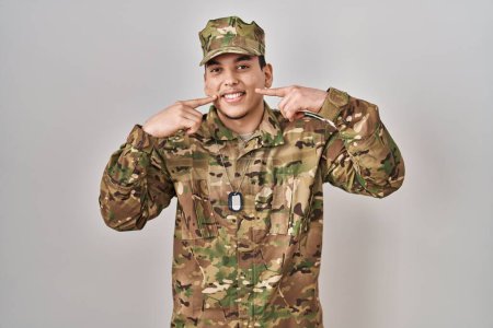 Téléchargez les photos : Jeune homme arabe portant uniforme de camouflage de l'armée souriant gai montrant et pointant du doigt les dents et la bouche. concept de santé dentaire. - en image libre de droit