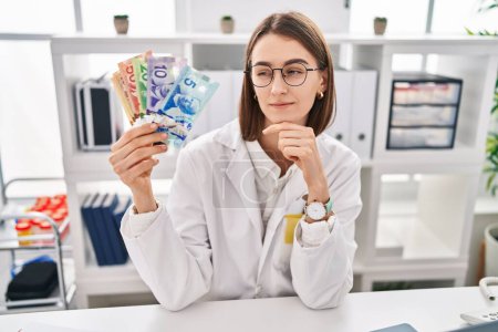Photo for Young caucasian doctor woman holding canadian dollars banknotes serious face thinking about question with hand on chin, thoughtful about confusing idea - Royalty Free Image