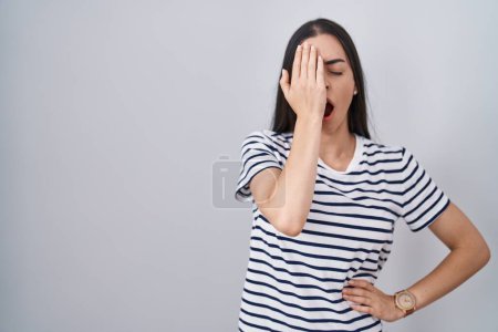 Téléchargez les photos : Jeune femme brune portant un t-shirt rayé bâillant fatigué couvrant la moitié du visage, des yeux et de la bouche avec la main. mal au visage dans la douleur. - en image libre de droit