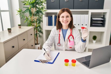 Photo for Young blonde woman doctor writing medical report holding empty test tube at clinic - Royalty Free Image