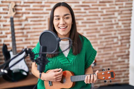 Foto de Joven mujer hispana músico cantando canción tocando ukelele en el estudio de música - Imagen libre de derechos