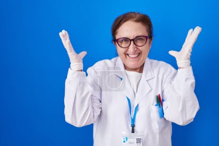 Foto de Mujer mayor con gafas con uniforme científico celebrando loco y loco por el éxito con los brazos levantados y los ojos cerrados gritando emocionado. concepto ganador - Imagen libre de derechos