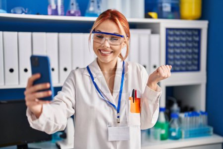 Photo for Young caucasian woman working at scientist laboratory doing video call with smartphone screaming proud, celebrating victory and success very excited with raised arm - Royalty Free Image