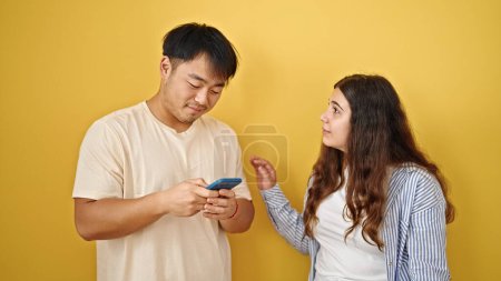 Téléchargez les photos : Couple homme et femme utilisant des smartphones avec une expression sérieuse sur fond jaune isolé - en image libre de droit
