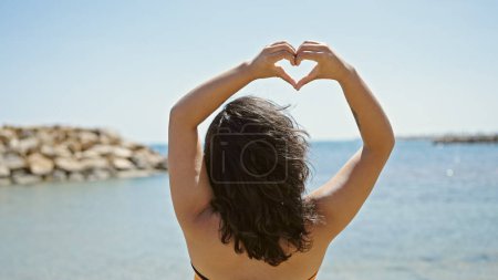Téléchargez les photos : Jeune belle hispanique femme touriste faire geste de coeur debout vers l'arrière à la plage - en image libre de droit