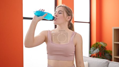 Foto de Mujer rubia joven que usa ropa deportiva bebiendo bebida energética en casa - Imagen libre de derechos