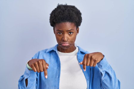 Foto de Mujer afroamericana de pie sobre fondo azul apuntando hacia abajo mirando triste y molesto, indicando la dirección con los dedos, infeliz y deprimido. - Imagen libre de derechos