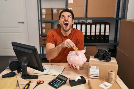 Photo for Young hispanic man working at small business ecommerce holding piggy bank angry and mad screaming frustrated and furious, shouting with anger looking up. - Royalty Free Image