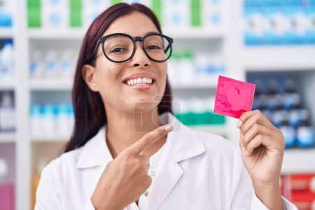 Téléchargez les photos : Jeune femme hispanique travaillant à la pharmacie tenant préservatif souriant heureux pointant avec la main et le doigt - en image libre de droit