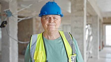Foto de Constructor de hombre de pelo gris de mediana edad sonriendo confiado de pie en el sitio de construcción - Imagen libre de derechos