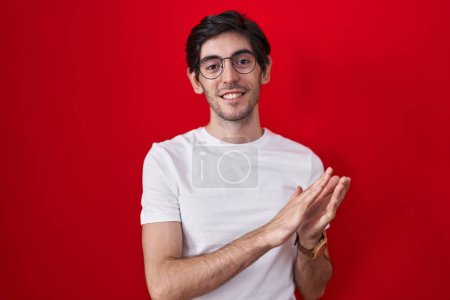 Téléchargez les photos : Jeune homme hispanique debout sur fond rouge applaudissant et applaudissant heureux et joyeux, souriant mains fières ensemble - en image libre de droit