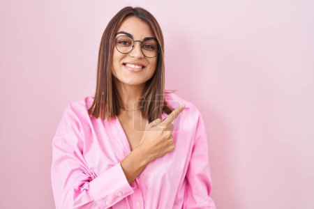Téléchargez les photos : Jeune femme hispanique portant des lunettes debout sur fond rose gai avec un sourire sur le visage pointant avec la main et le doigt vers le côté avec une expression heureuse et naturelle - en image libre de droit