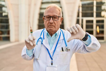 Photo for Senior doctor with grey hair holding syringe with angry face, negative sign showing dislike with thumbs down, rejection concept - Royalty Free Image