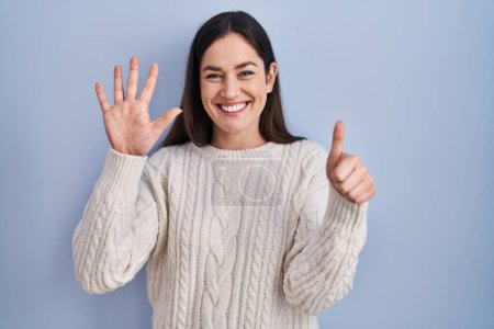Téléchargez les photos : Jeune femme brune debout sur fond bleu montrant et pointant vers le haut avec les doigts numéro six tout en souriant confiant et heureux. - en image libre de droit
