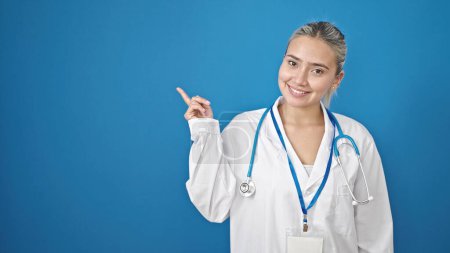 Foto de Joven hermosa mujer hispana médico sonriendo señalando a un lado sobre aislado fondo azul - Imagen libre de derechos
