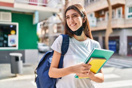 Foto de Joven hermosa estudiante hispana con máscara médica sosteniendo libros en la calle - Imagen libre de derechos