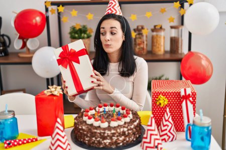 Foto de Hispanic woman celebrating birthday with cake holding present smiling looking to the side and staring away thinking. - Imagen libre de derechos