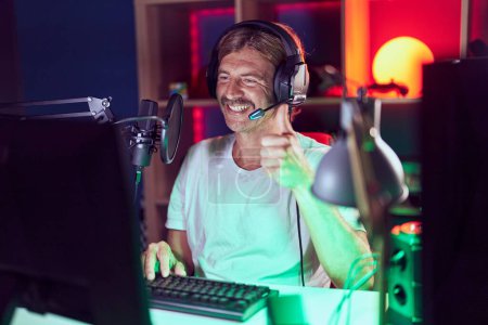 Téléchargez les photos : Homme caucasien avec moustache jouer à des jeux vidéo souriant heureux et positif, pouce levé faire excellent et signe d'approbation - en image libre de droit