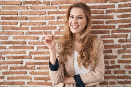 Photo for Beautiful blonde woman standing over bricks wall with a big smile on face, pointing with hand and finger to the side looking at the camera. - Royalty Free Image