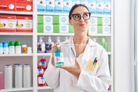 Téléchargez les photos : Femme brune travaillant à la pharmacie drugstore tenant sirop contre la toux souriant regardant vers le côté et regardant loin en pensant. - en image libre de droit