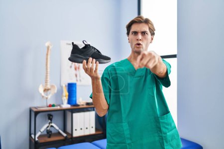 Téléchargez les photos : Jeune homme travaillant à la clinique de physiothérapie tenant la chaussure pointant du doigt vers la caméra et vers vous, geste confiant et sérieux - en image libre de droit