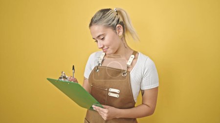 Téléchargez les photos : Jeune belle femme hispanique portant tablier écriture sur presse-papiers sur fond jaune isolé - en image libre de droit