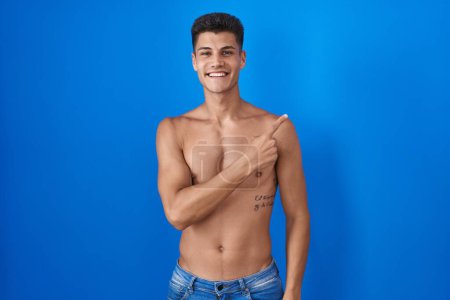 Foto de Joven hombre hispano de pie sin camisa sobre fondo azul alegre con una sonrisa en la cara señalando con la mano y el dedo hacia el lado con expresión feliz y natural - Imagen libre de derechos