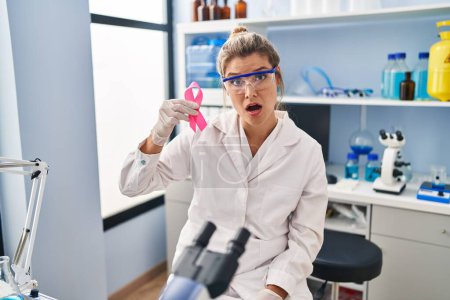 Foto de Mujer joven trabajando en laboratorio científico sosteniendo cinta rosa asustada y sorprendida con la boca abierta para sorpresa, cara de incredulidad - Imagen libre de derechos