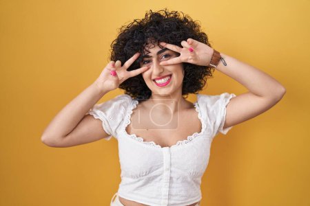 Téléchargez les photos : Jeune femme brune avec les cheveux bouclés debout sur fond jaune faisant symbole de paix avec les doigts sur le visage, souriant joyeuse montrant la victoire - en image libre de droit
