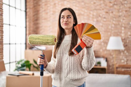 Photo for Young brunette woman holding roller painter and paint samples smiling looking to the side and staring away thinking. - Royalty Free Image