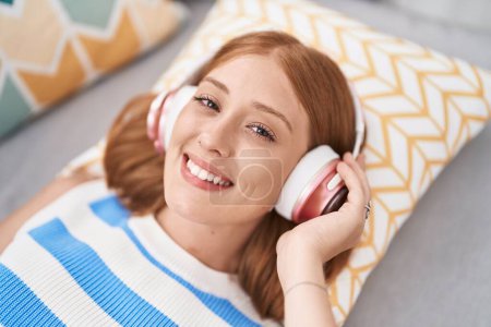 Young redhead woman listening to music lying on sofa at home