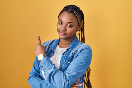 Photo for African american woman with braids standing over yellow background pointing with hand finger to the side showing advertisement, serious and calm face - Royalty Free Image