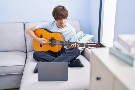 Foto de Joven hombre rubio que tiene clase de guitarra clásica en línea sentado en el sofá en casa - Imagen libre de derechos